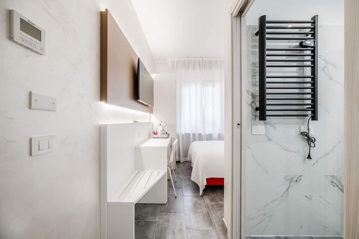A modern hotel room with a bed, a desk, a TV, and a bathroom with a shower and a towel rack.