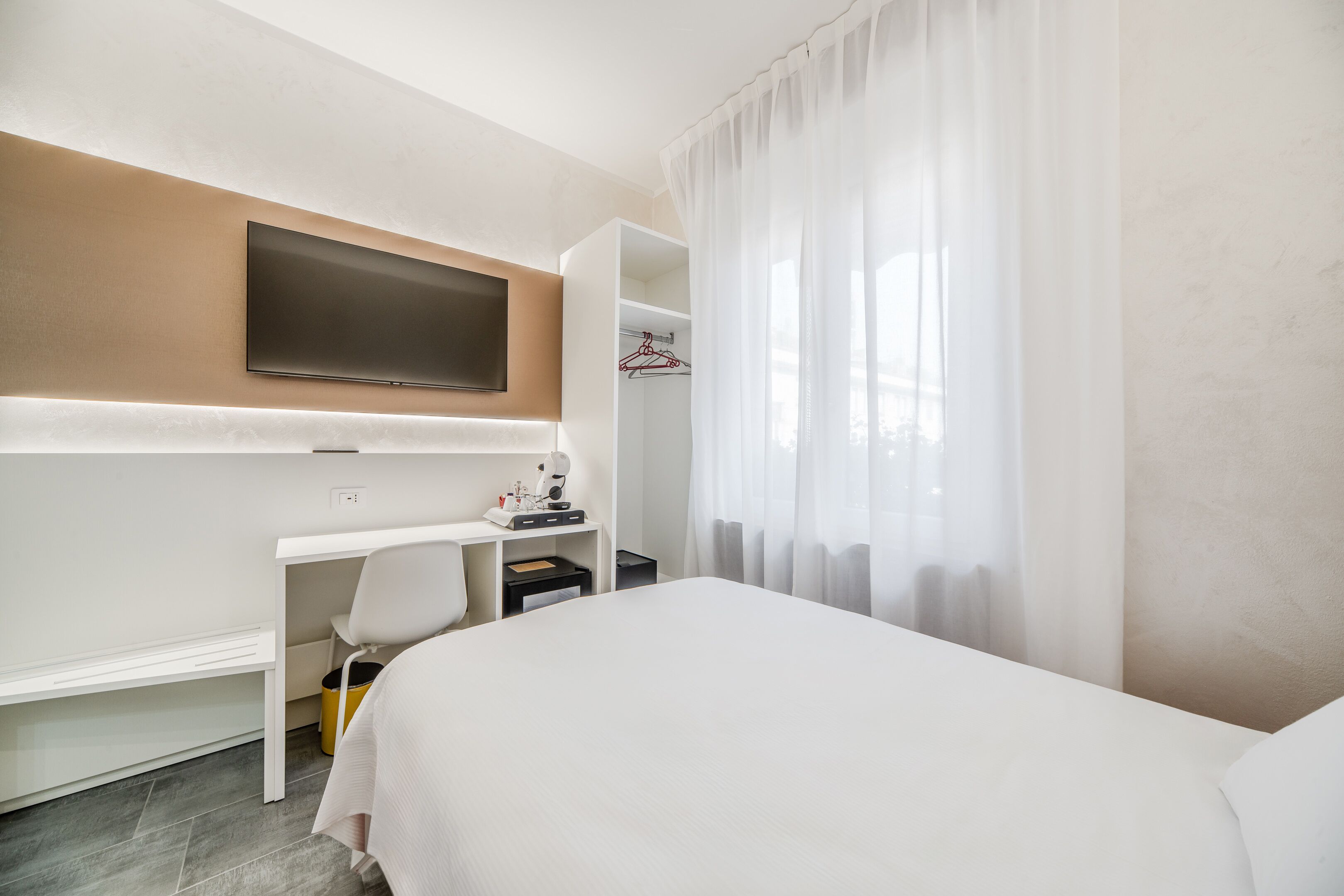 Una habitación de hotel con cama, escritorio, silla, televisor montado en la pared y armario.