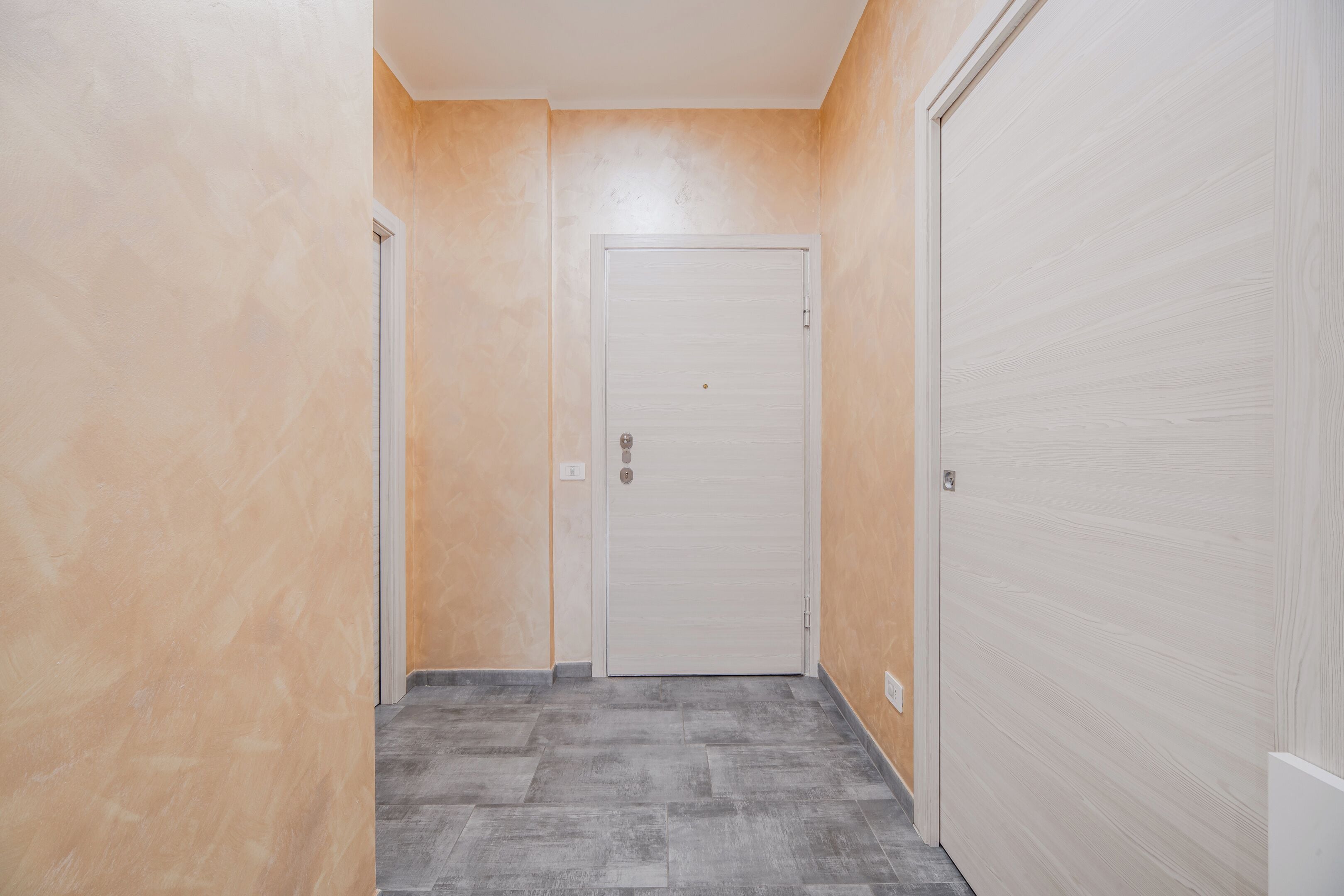 A hallway with a closed door, a light switch, and a power outlet on the wall.
