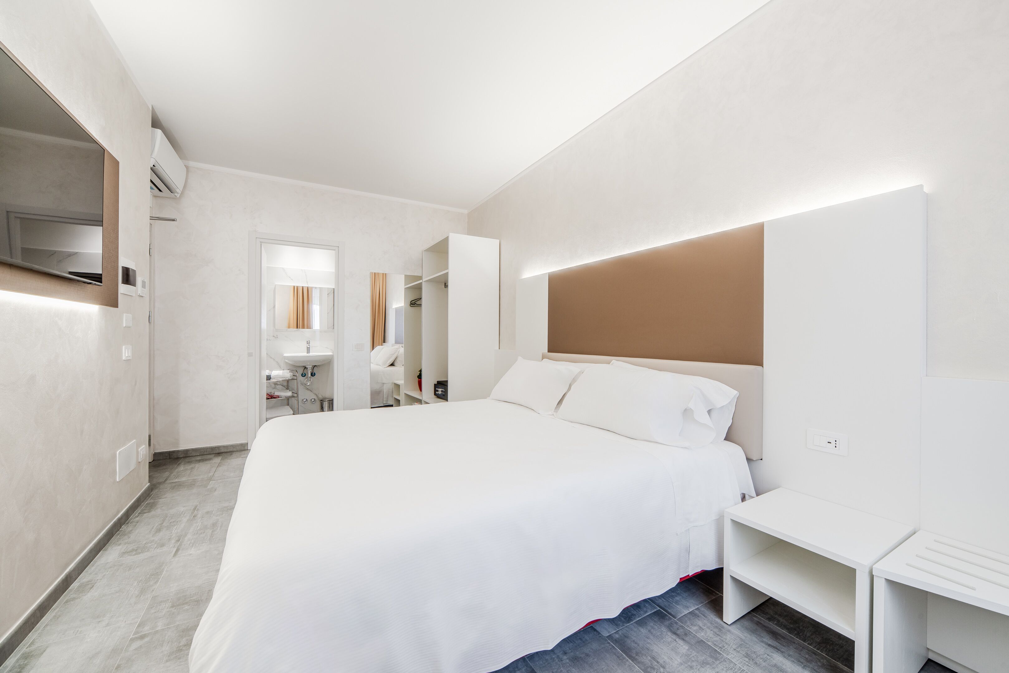 A modern hotel room with a large bed, bedside tables, a built-in TV, and a bathroom with a mirror and sink.