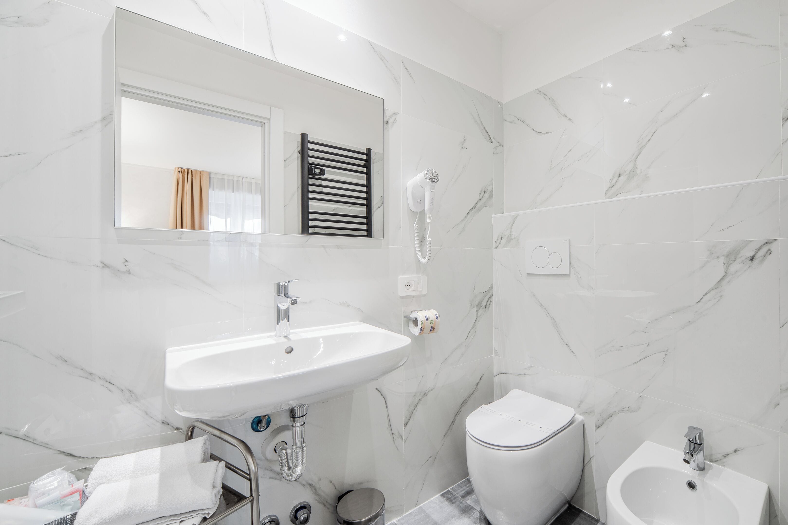 A modern bathroom with a white sink, toilet, and bidet, marble walls, and a towel rack.