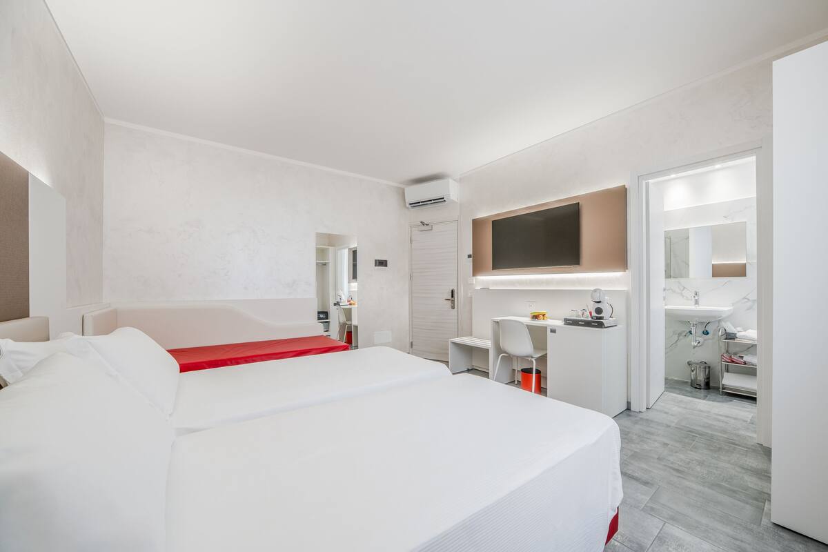 A modern hotel room with a large bed, a desk, a chair, a mirror, and a bathroom with a sink and mirror.