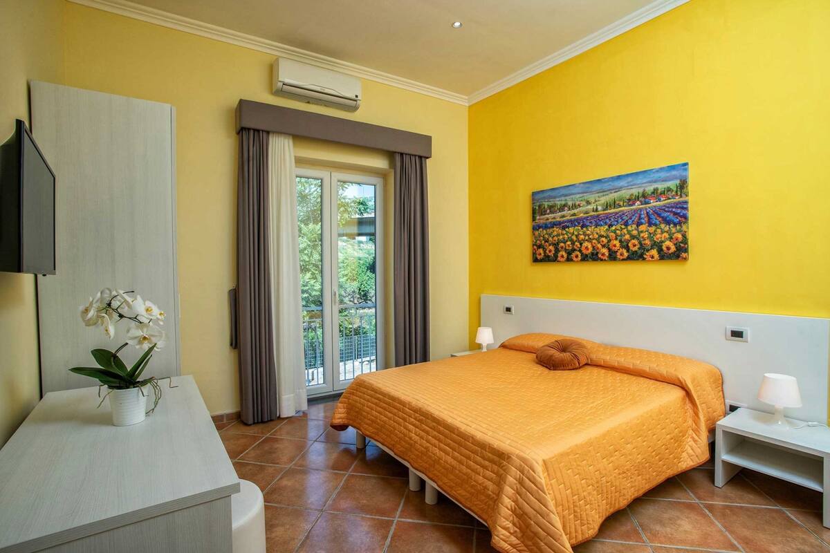 A bedroom with a bed, a desk with a plant, a painting on the wall, and a window with curtains.