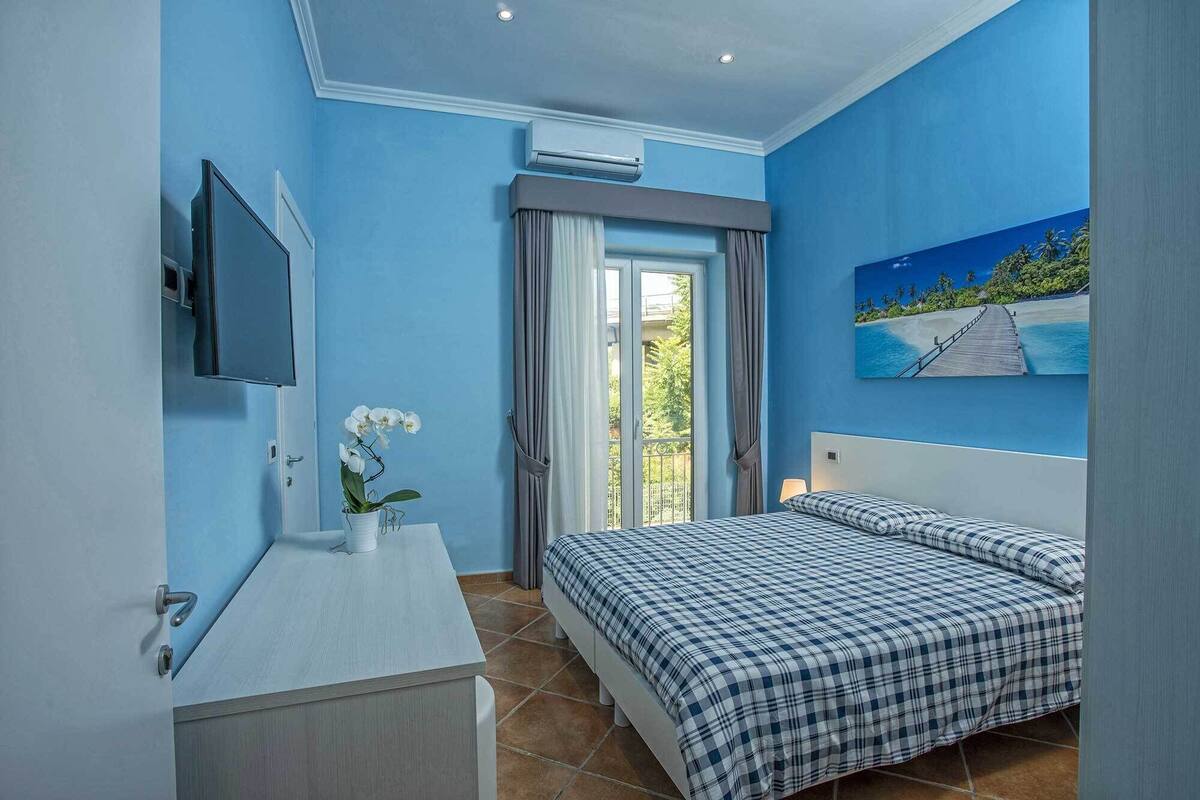 A bedroom with a bed, a TV, a window with curtains, and a painting of a beach scene.