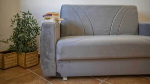 A grey sofa with a cushioned backrest, a small wooden side table with a plant and books, and a tiled floor. - B&B Carmelina (Gragnano)