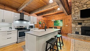 A modern kitchen with white cabinets, a central island, a stone fireplace, and wooden beams on the ceiling.