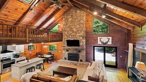 A spacious lodge with a stone fireplace, wooden beams, a balcony, and a kitchen area.