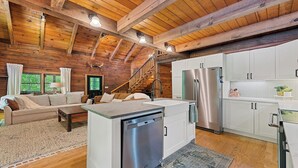 A cozy cabin interior with a wooden ceiling, a kitchen with stainless steel appliances, a dining area with a table and chairs, and a staircase leading to a loft.