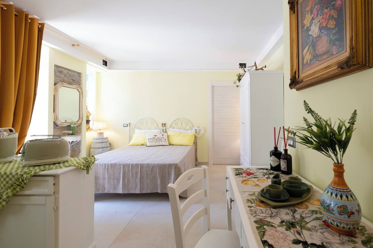 Un dormitorio con cama, mesita de noche y una silla. Hay una ventana con cortinas, un espejo y un cuadro en la pared.