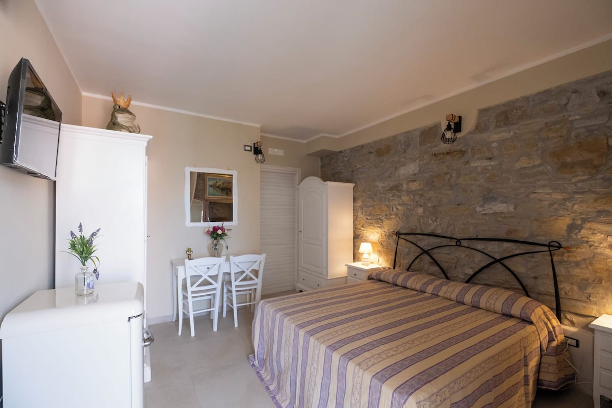 Un dormitorio con pared de piedra, una cama con edredón a rayas, un escritorio con silla y una mesita de noche con lámpara.