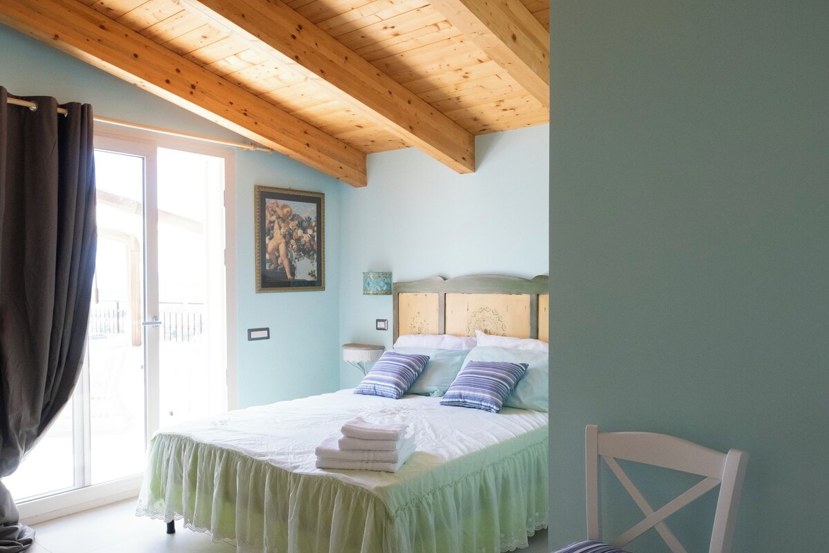 A bedroom with a wooden ceiling, a bed with white bedding, a chair with a striped cushion, and a painting on the wall.