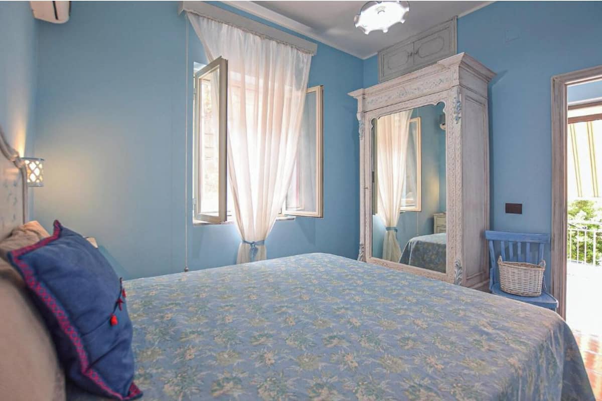 Un dormitorio con cama, mesita de noche, espejo y ventana con cortinas.