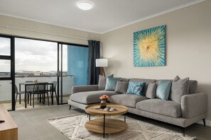 A modern living room with a grey sofa, a round coffee table, and a large window with a view. - 2 Bed Apartment in Century City | Pool (Cape Town)