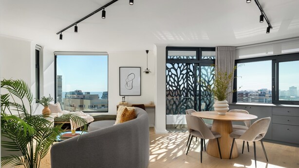 Un salon moderne avec un canapé gris aux lignes courbes, une table de salle à manger ronde en bois entourée de chaises, et de grandes fenêtres offrant une vue sur la ville.