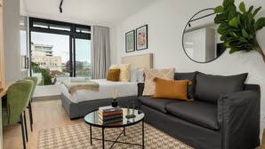 A modern hotel room with a bed, a sofa, a round coffee table, and a large window offering a city view. - The Sage l Studio l Luxury Finishes and Balcony View (Cape Town)