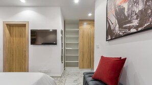 A modern bedroom with a bed, a grey sofa with a red pillow, a TV mounted on the wall, and a wooden wardrobe. - De Castle hotels & Resort (Enugu)