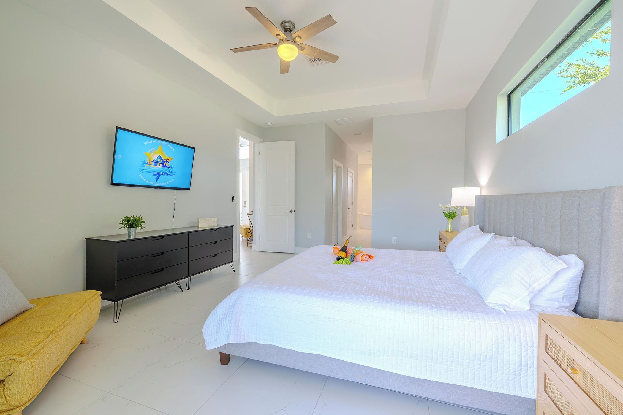 A bedroom with a bed, a ceiling fan, a TV, a dresser, and a window.