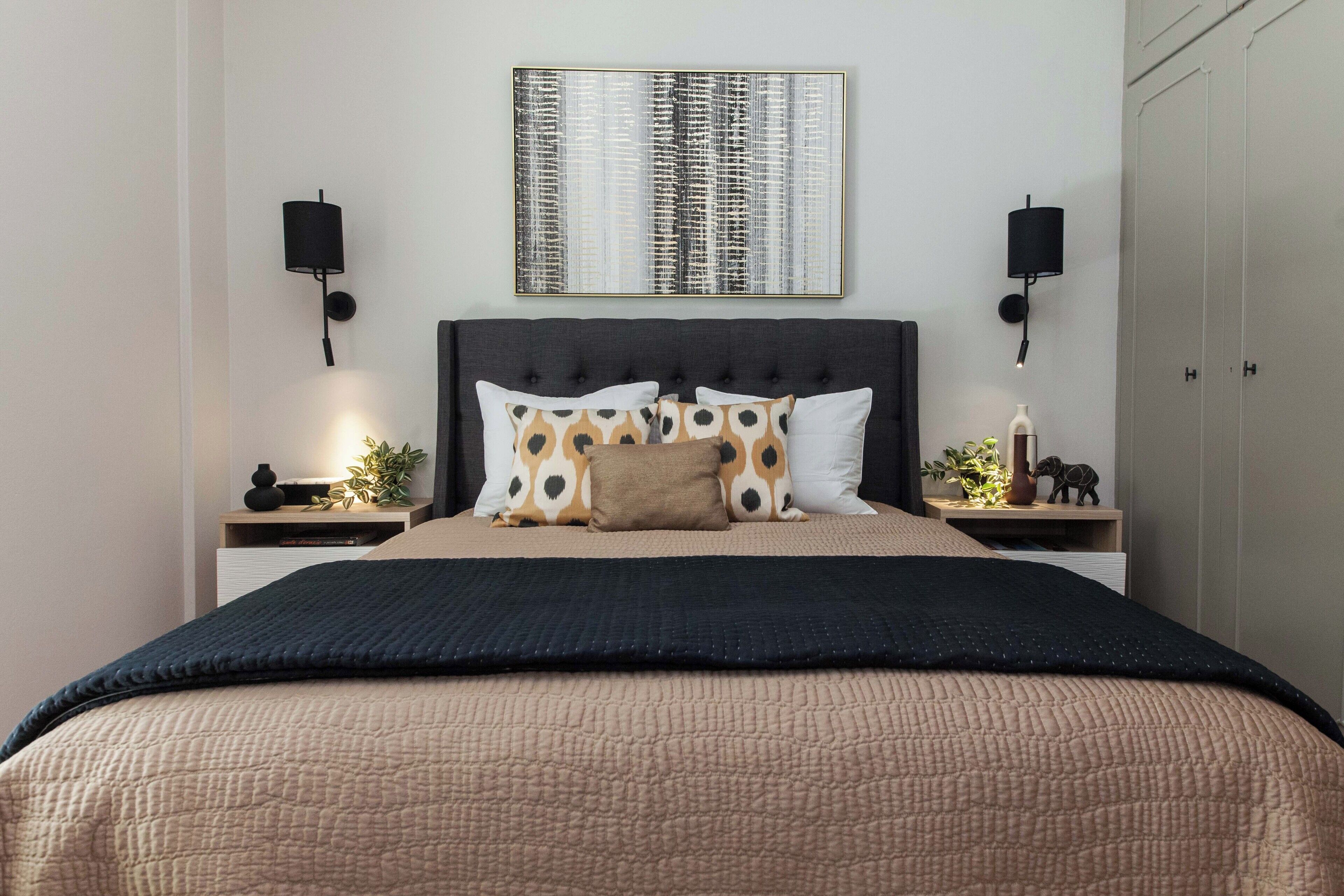 Una cama bien tendida con almohadas estampadas, un cabecero oscuro y dos mesitas de noche con lámparas y objetos decorativos.