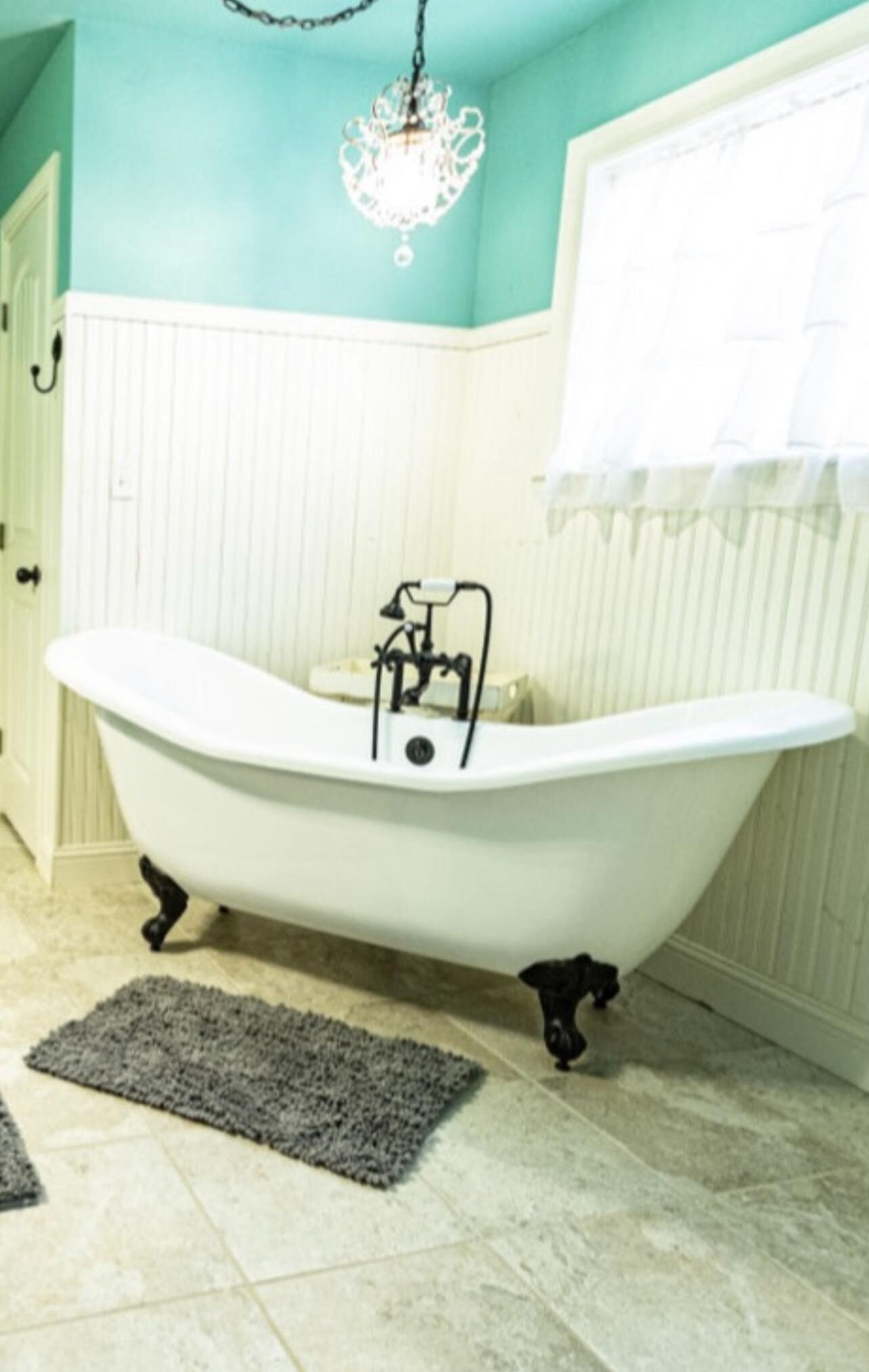 Une salle de bain avec une baignoire sur pieds, un lustre, une fenêtre avec un rideau blanc et un sol carrelé.