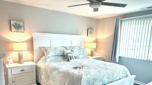 Une chambre à coucher bien rangée, avec un lit, deux tables de chevet, un ventilateur de plafond et une fenêtre avec des stores.