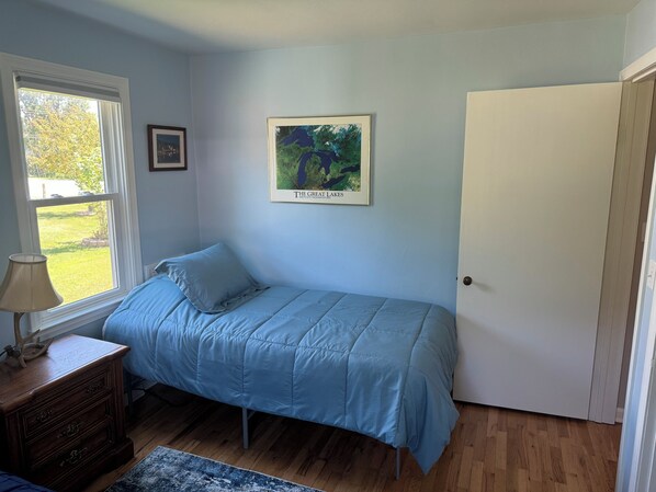 A bedroom with a single bed, a window with a view of a grassy yard, a framed map on the wall, and a closed door.