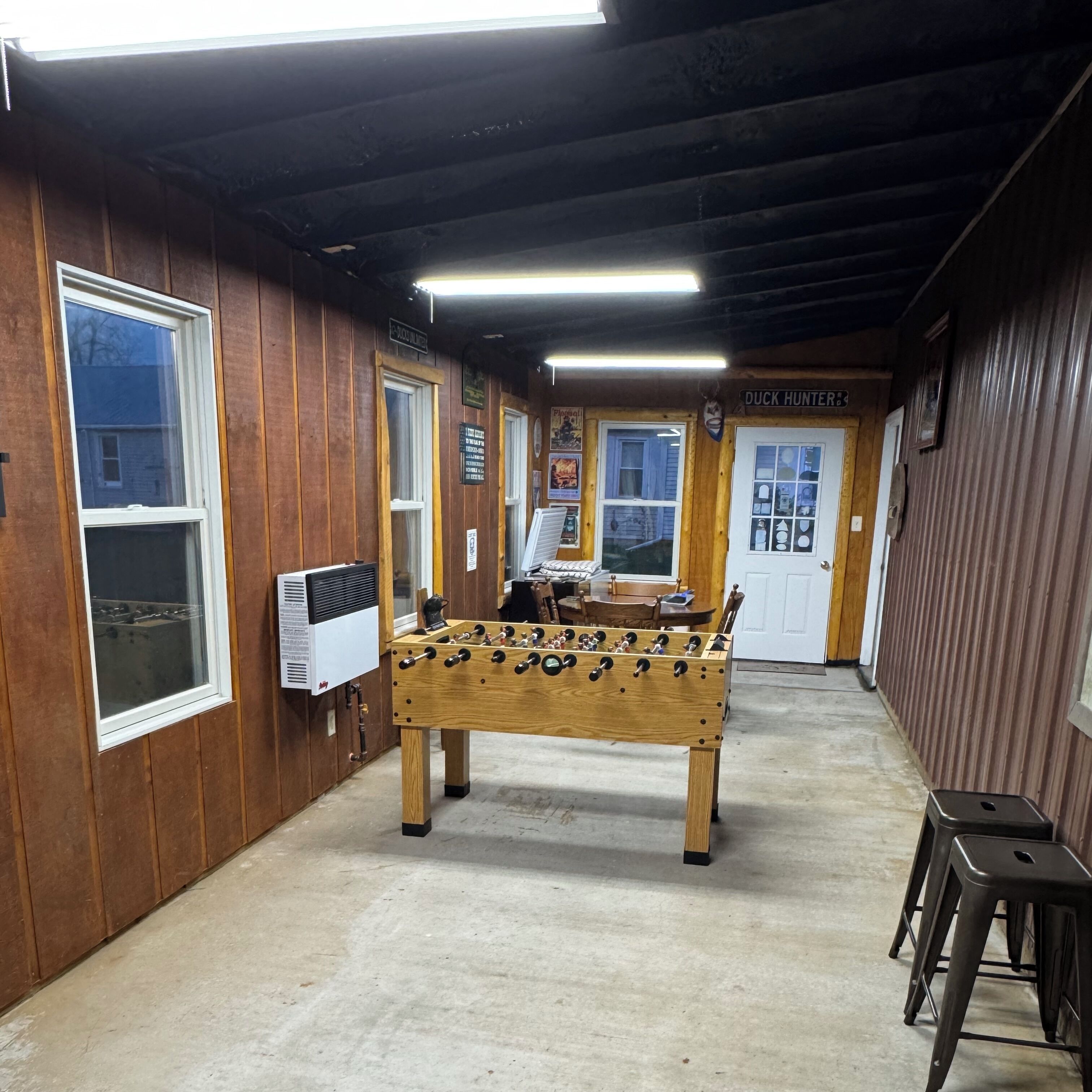 Quarto com piso de madeira, uma mesa de pebolim, uma porta com a inscrição "Duck Hunter" e uma janela com vista para uma casa.