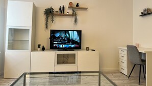 Una sala moderna con un televisor de pantalla plana montado en un mueble blanco, una mesa de centro de vidrio, una silla de oficina gris y un escritorio blanco con una silla.