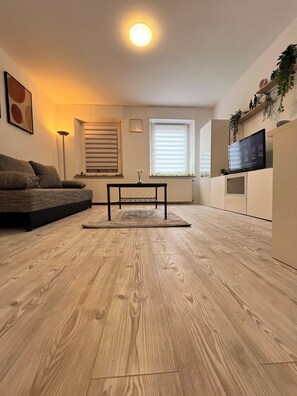 Un salon moderne avec un plancher en bois, un canapé, une table basse, un téléviseur posé sur un meuble et une fenêtre avec des stores.