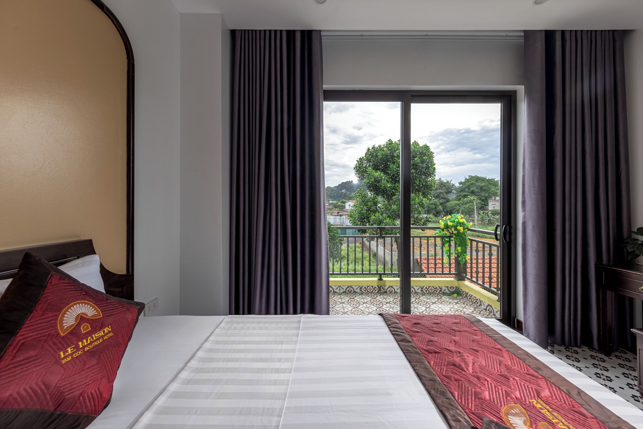 Ein Hotelzimmer mit Bett, Blick auf einen Balkon mit Geländer und Bepflanzung sowie ein Fenster mit Vorhängen.