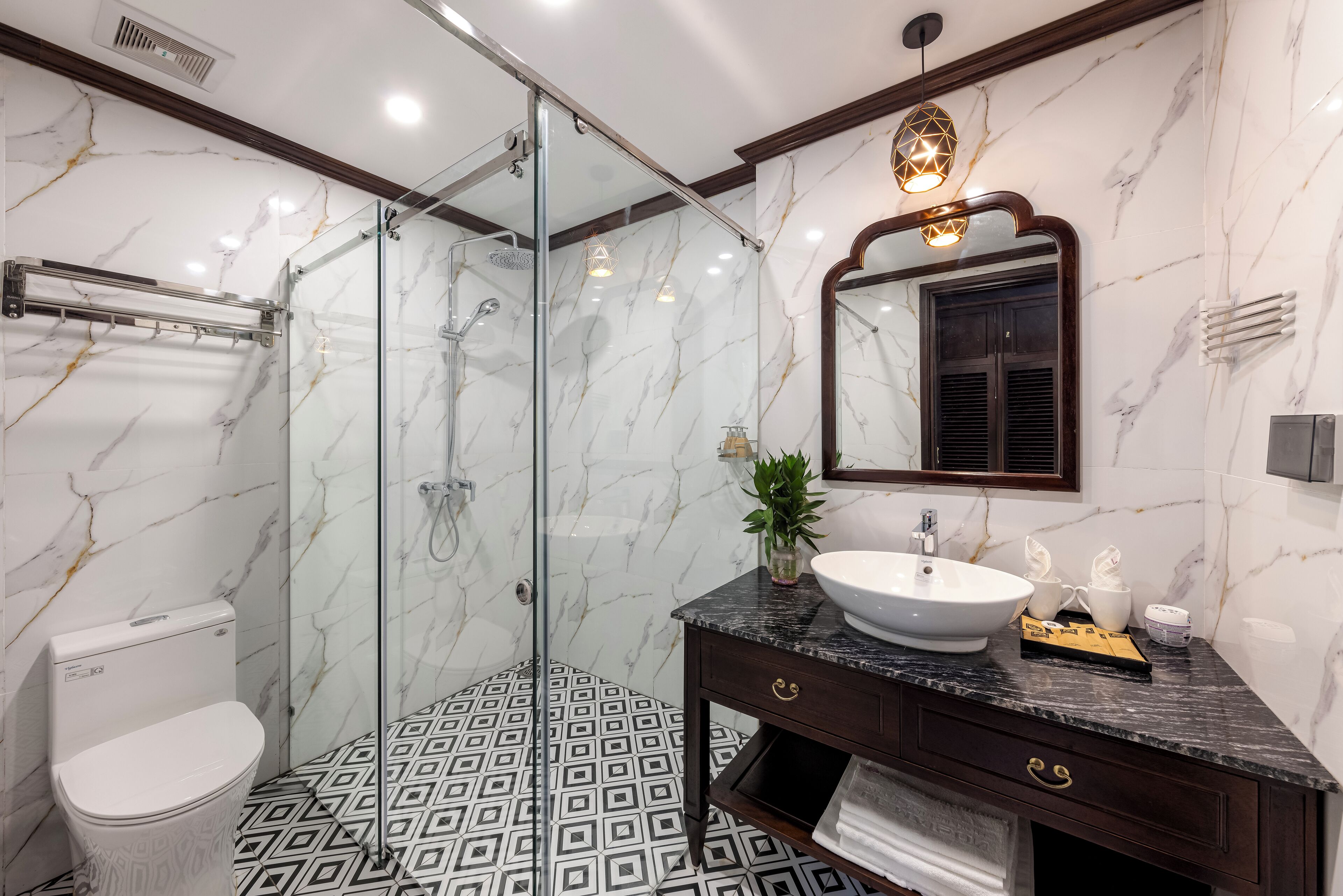 Un baño moderno con ducha envuelta en vidrio, inodoro blanco, piso con diseño de mármol y tocador de mármol oscuro con lavamanos y espejo.