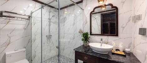 Une salle de bain moderne avec une douche à parois de verre, des toilettes blanches, un carrelage imitant le marbre et un meuble-lavabo en marbre foncé, doté d’un évier et d’un miroir.