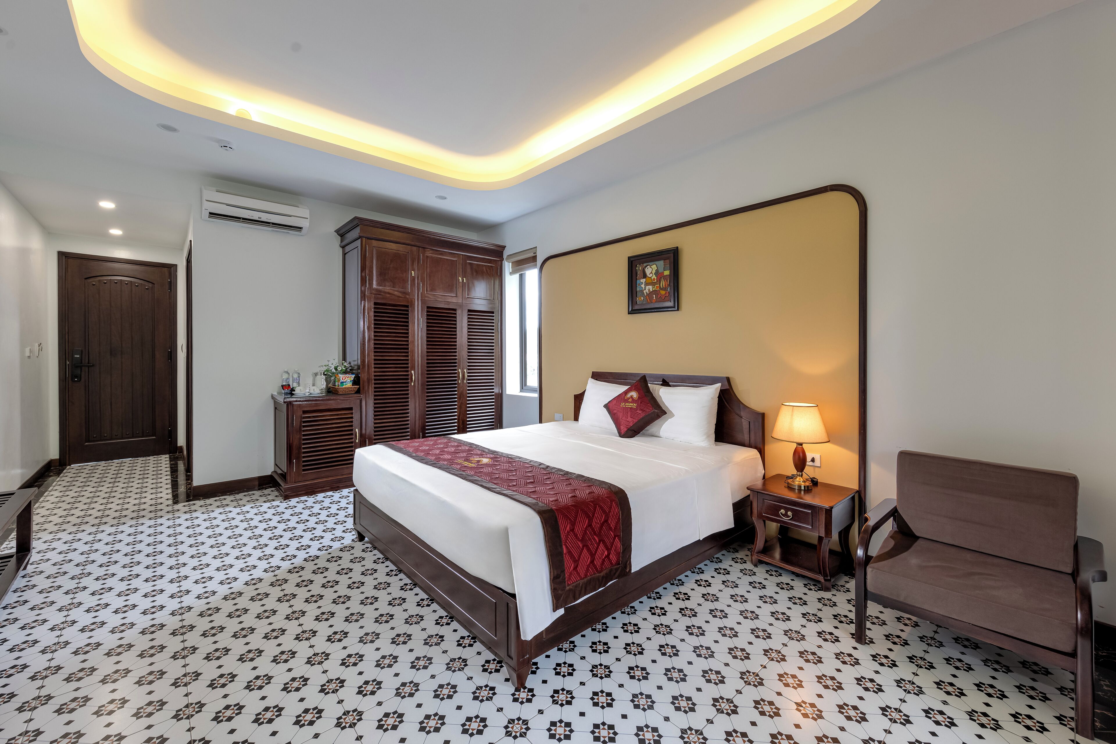 Habitación de hotel con una cama grande, una mesita de noche con lámpara, una silla, un armario y baldosas de suelo estampadas.