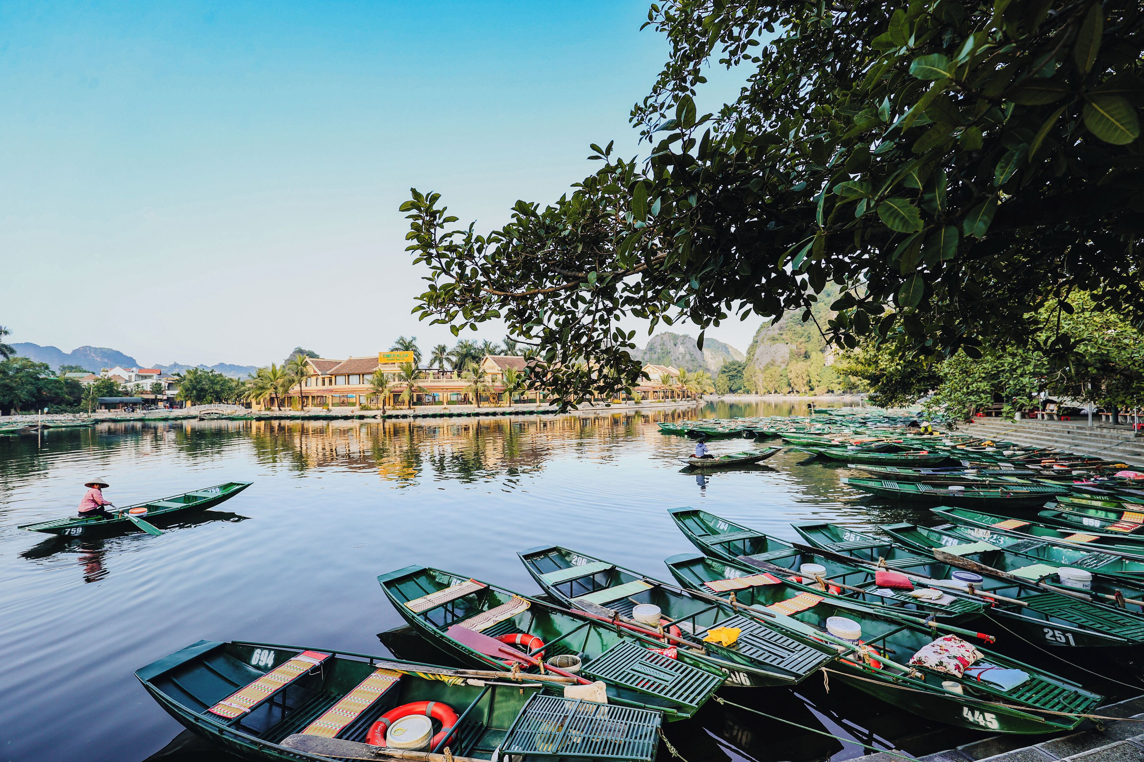 A calm river with numerous green boats docked along the shore, a bridge in the background, and buildings with traditional architecture.