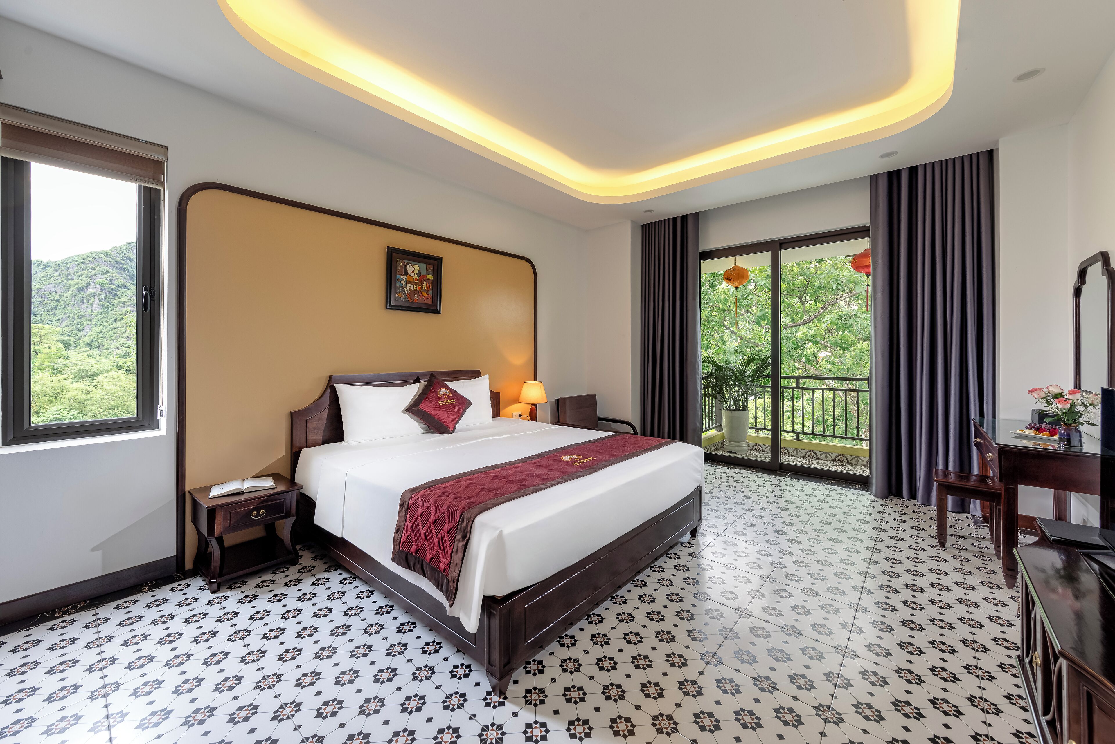 A hotel room with a large bed, a nightstand, a desk, and a view of greenery through the window.