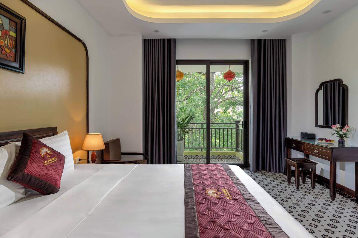 A hotel room with a bed, a desk, a chair, a lamp, and a view of greenery outside.