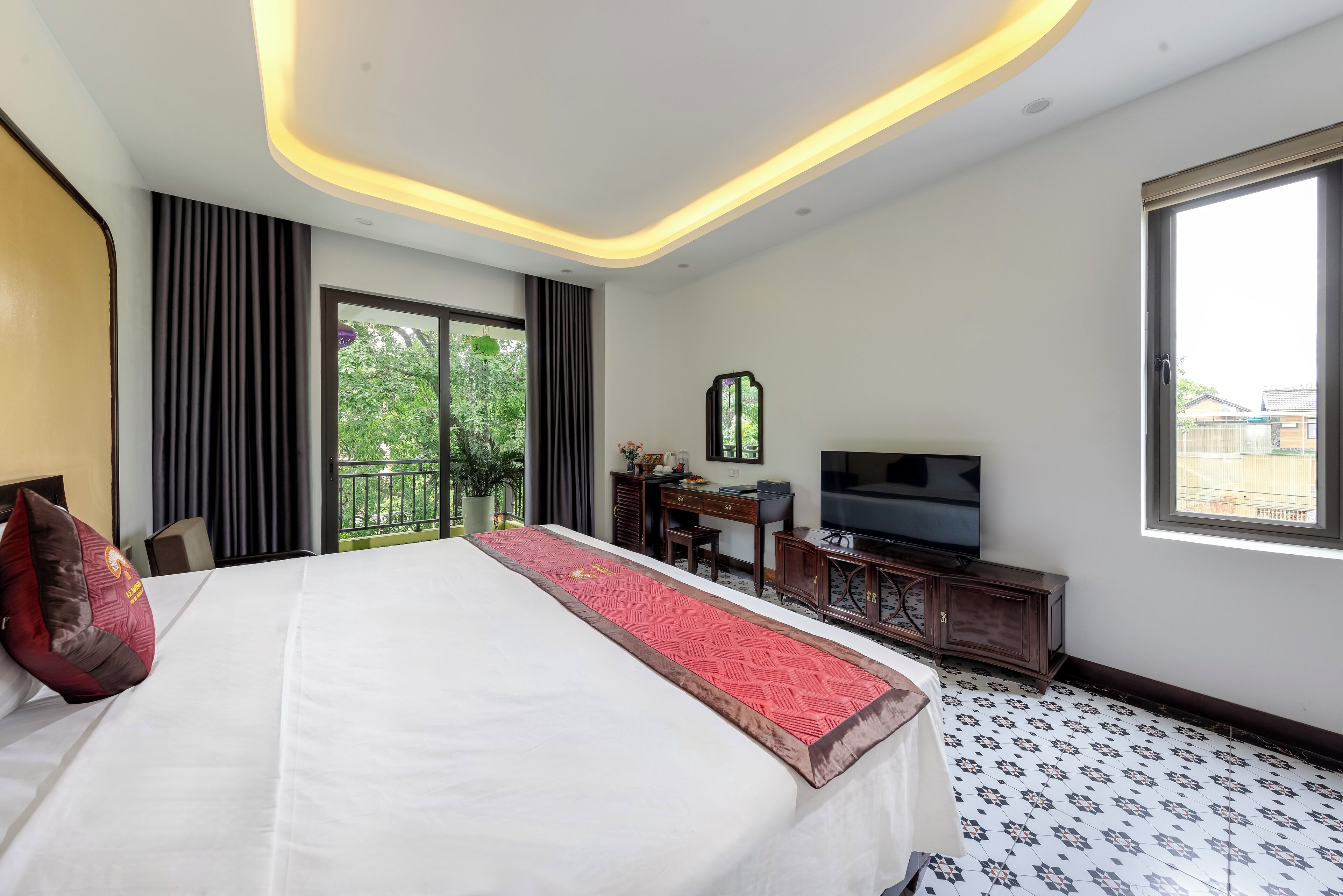A hotel room with a large bed, a desk with a television, and a view of greenery outside.