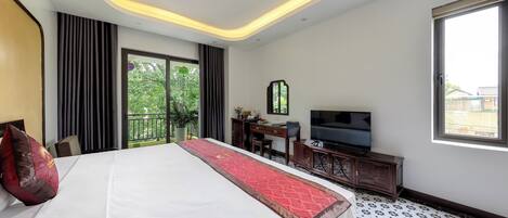 A hotel room with a large bed, a desk with a television, and a view of greenery outside.