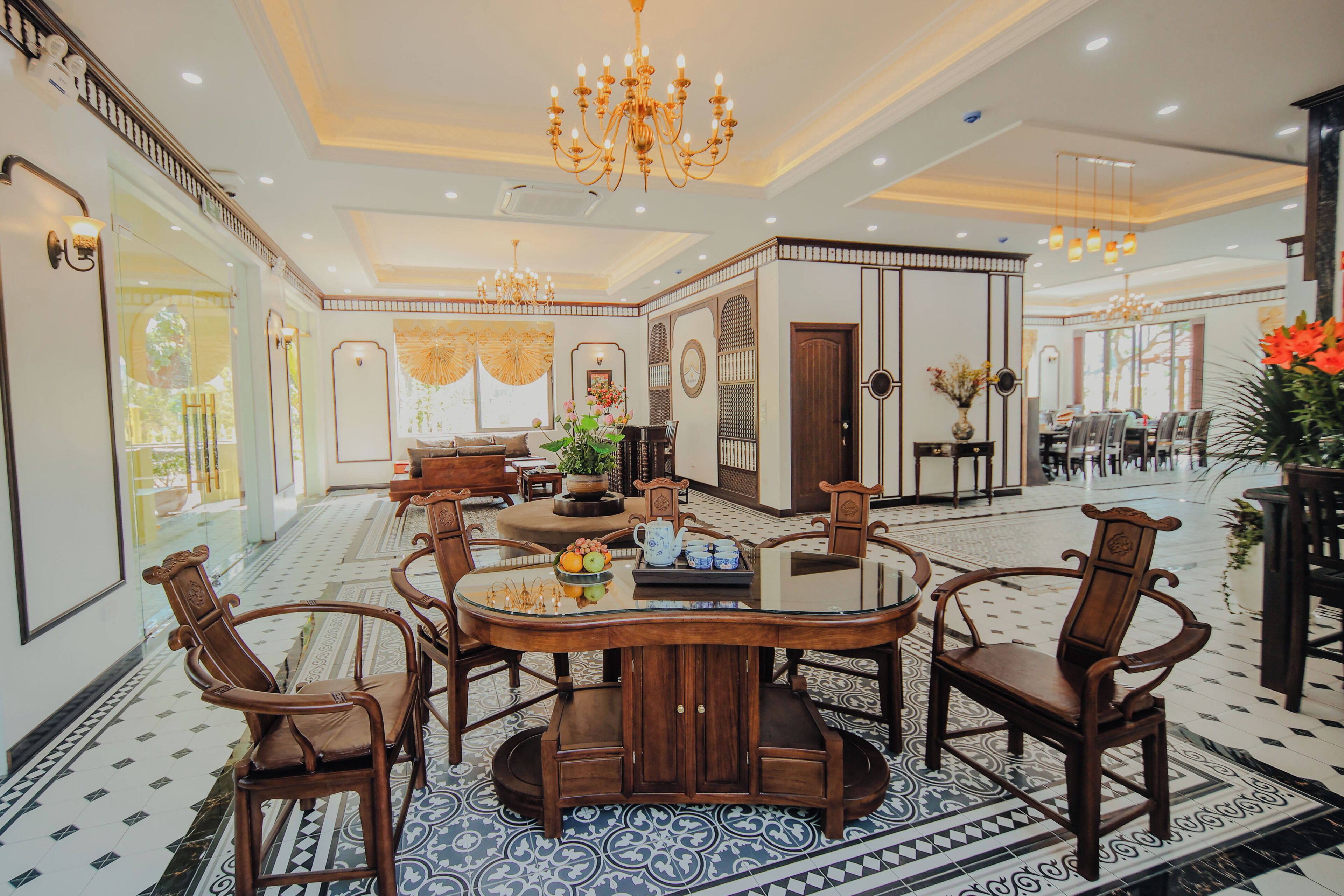 A spacious hotel lobby with a round dining table surrounded by chairs, a chandelier, and a patterned floor.