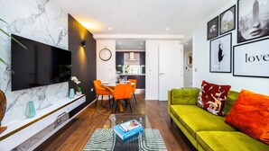 A modern living room with a flat-screen TV, a green sofa, and a dining area with orange chairs. - Cosy Corner Apartment Near to Excel (London)