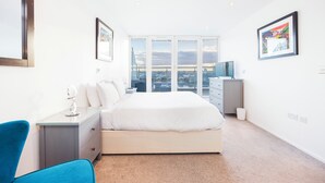 A modern bedroom with a large bed, a desk with a mirror, and a view of the city through large windows. - Cosy Corner Apartment Near to Excel (London)