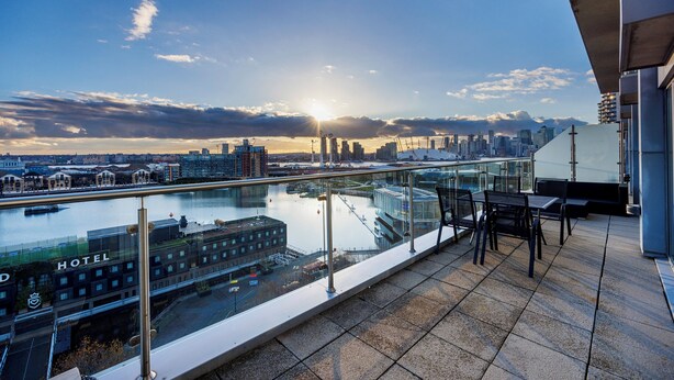 A rooftop terrace with a glass balustrade overlooking a cityscape and a body of water. - Cosy Corner Apartment Near to Excel (London)