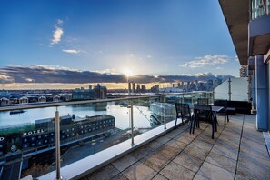 Eine Dachterrasse mit Glasbrüstung und Blick auf eine Skyline sowie ein Gewässer.