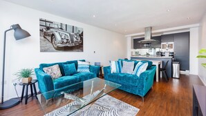 A modern living room with teal sofas, a glass coffee table, and a kitchen in the background. - Cosy Corner Apartment Near to Excel (London)