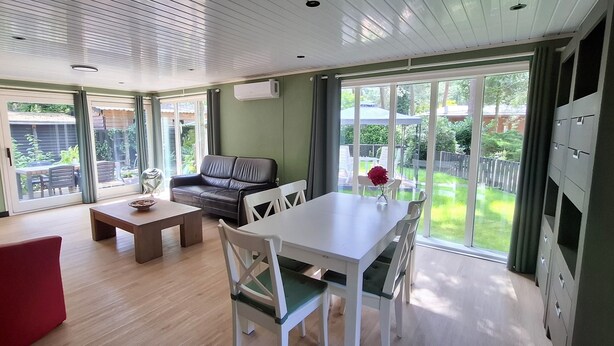 A modern living room with a sofa, a dining table, and chairs. There is a large window with a view of a garden.