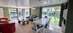 A modern living room with a sofa, a dining table, and chairs. There is a large window with a view of a garden.