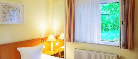 A hotel room with a bed, a nightstand, a lamp, a window with curtains, and a radiator.