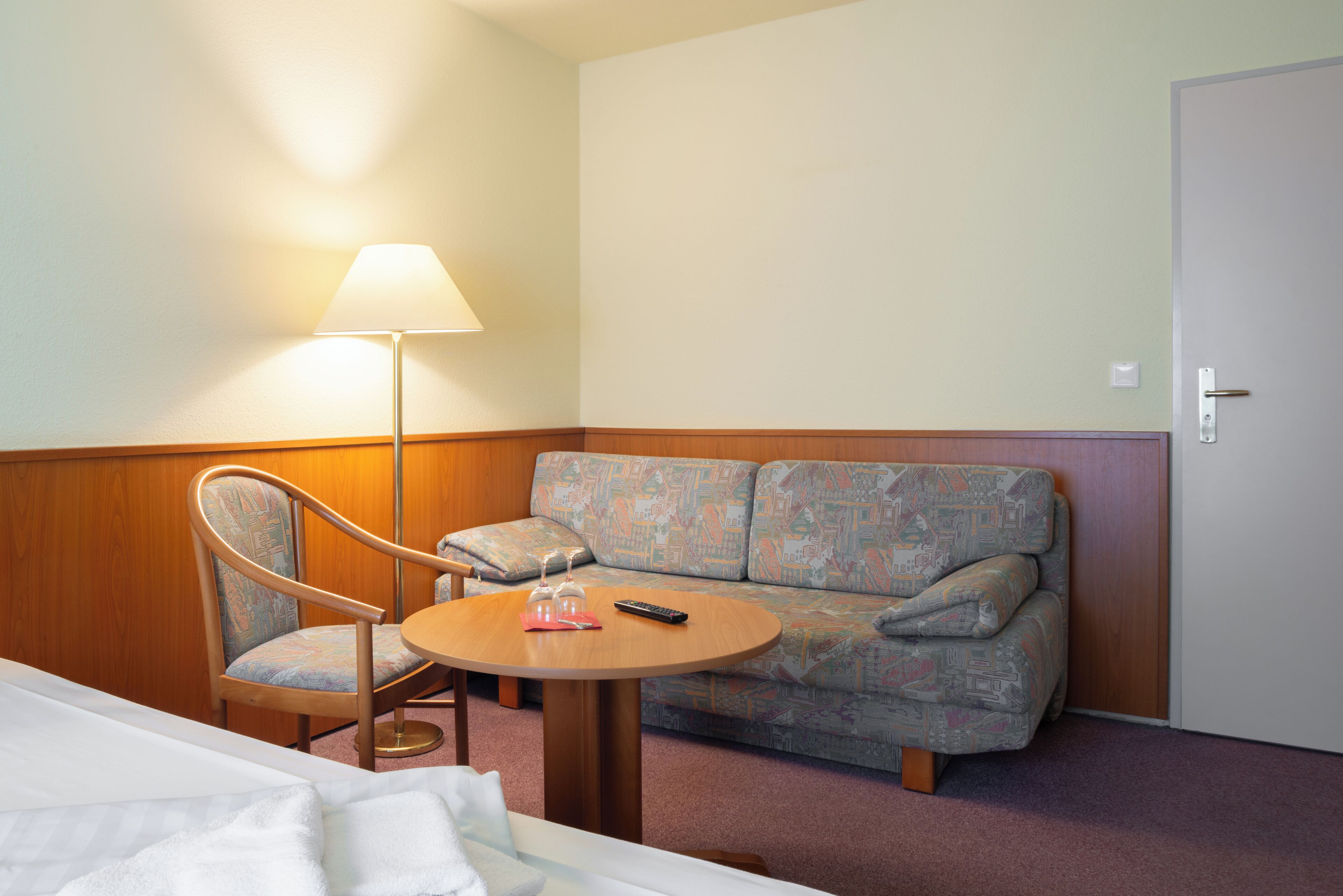 Una habitación de hotel con cama, una silla, un sofá, una mesita y una lámpara.