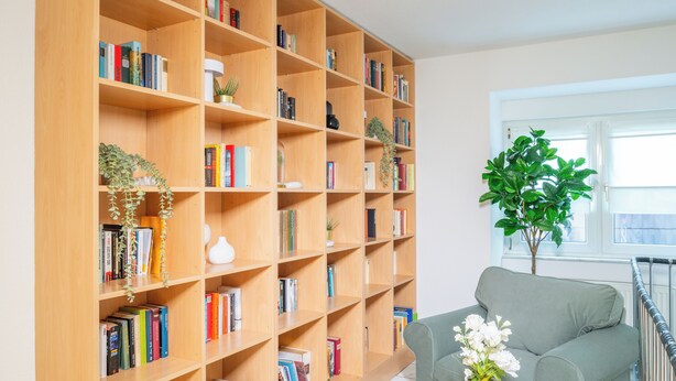 A living room with a large wooden bookshelf, a grey sofa, and a glass coffee table with a woven rug. - Waldhotel Schöneiche - Self-Check-in (Schoeneiche bei Berlin)