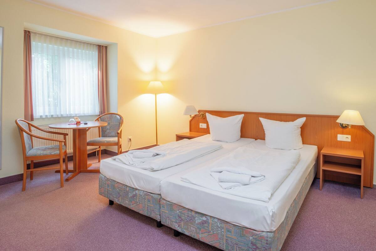 A hotel room with a double bed, a chair, a small table, a lamp, and a window with curtains.
