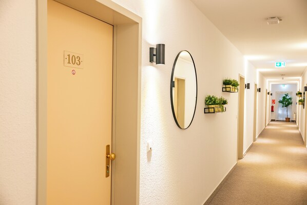 A hotel hallway with a door labeled '103', a wall-mounted mirror, and potted plants. - Waldhotel Schöneiche - Self-Check-in (Schoeneiche bei Berlin)